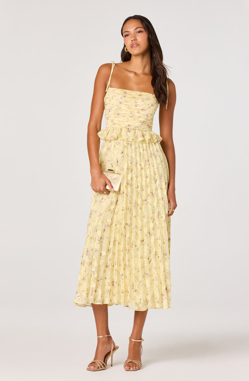 Woman in a yellow floral pleated dress with spaghetti straps, holding a clutch.