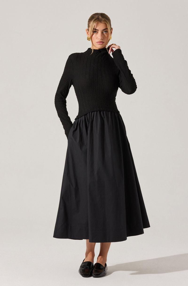 A woman is wearing a fitted black sweater with a textured, ribbed pattern and long sleeves. She pairs it with a high-waisted, full-length black skirt that has a gathered waistband, creating a flowing silhouette. She completes her outfit with black slip-on shoes.