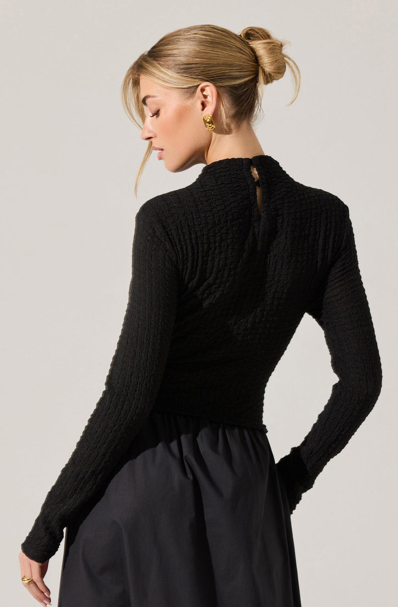 A woman is wearing a fitted, long-sleeved black sweater with a textured, chunky knit pattern. The sweater has a small opening at the back near the neckline, closed with a button. She pairs it with a high-waisted, black skirt made of a shiny, slightly puffy fabric. She accessorizes with large, gold earrings. Her hair is styled in a low, messy bun.