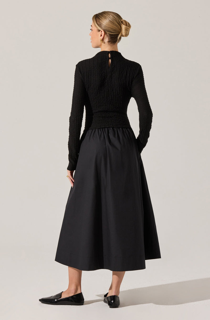 A woman is wearing a long black pleated skirt that reaches her mid-calf, paired with a textured black long-sleeve top with a keyhole opening at the back. She completes her outfit with black flat shoes.