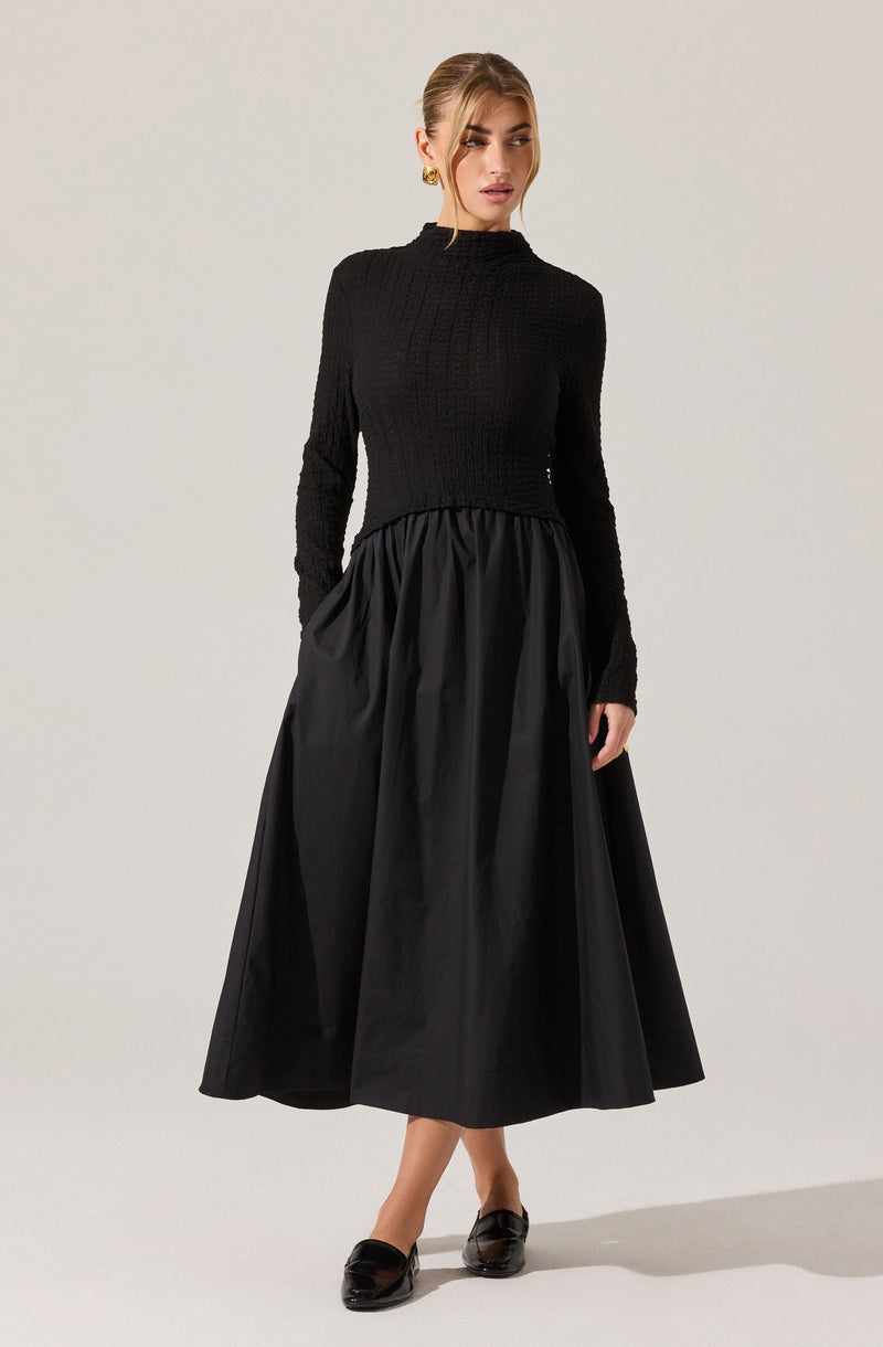 A woman is wearing a long-sleeved, fitted black sweater with a ribbed texture, paired with a high-waisted, full-length black skirt made of a shiny, gathered fabric. She has on black loafers and accessorizes with gold hoop earrings.