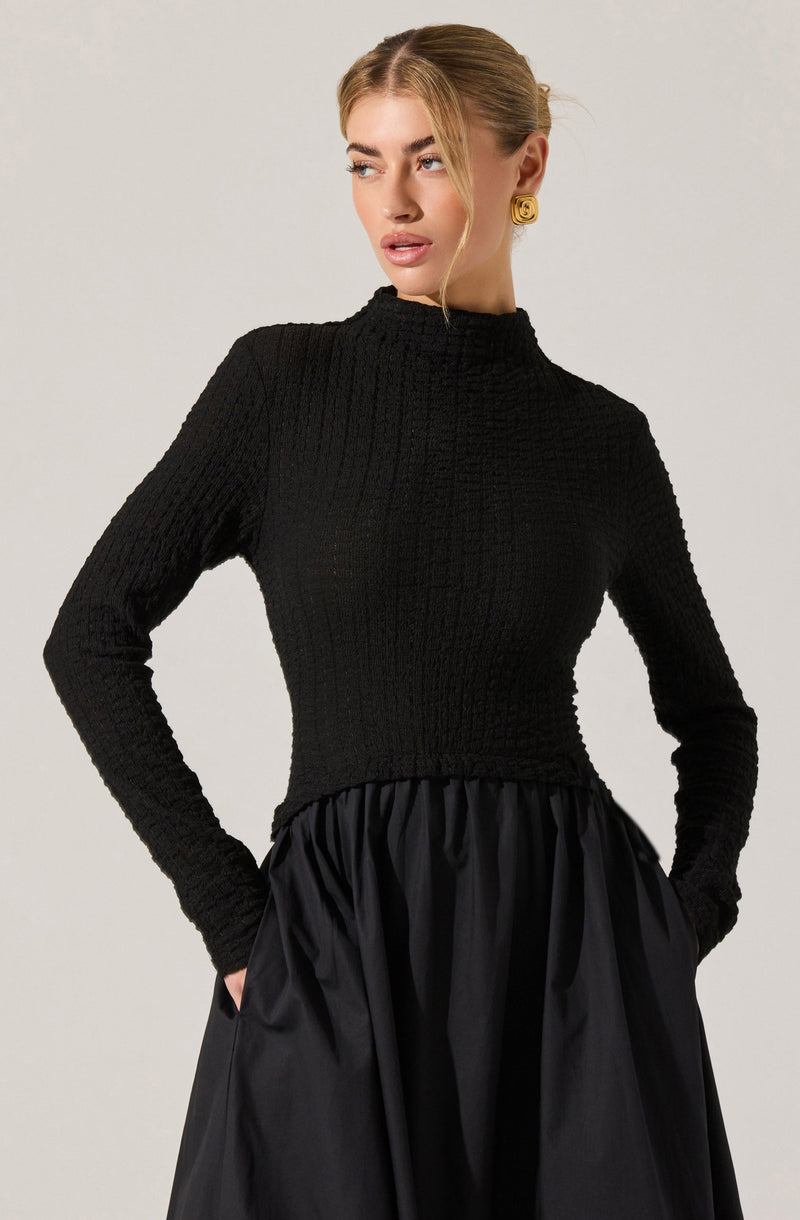 A woman is wearing a fitted, long-sleeve black ribbed sweater with a high neckline. She has a gold spiral earring in one ear and her hair is styled in a sleek, pulled-back look. The skirt she is wearing is black, full, and gathered, with a shiny, satin-like texture.