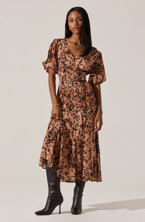 A woman is wearing a long, flowy dress with a leopard print pattern in shades of tan and black. The dress has puffed short sleeves and a V-neckline. She has paired the dress with tall black heeled boots and is accessorizing with a gold bracelet.