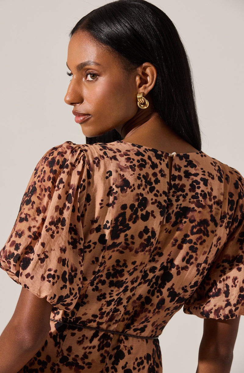A woman is wearing a dress with a leopard print pattern in shades of brown and black. The dress has short, puffy sleeves and a fitted waist. She accessorizes with gold hoop earrings.