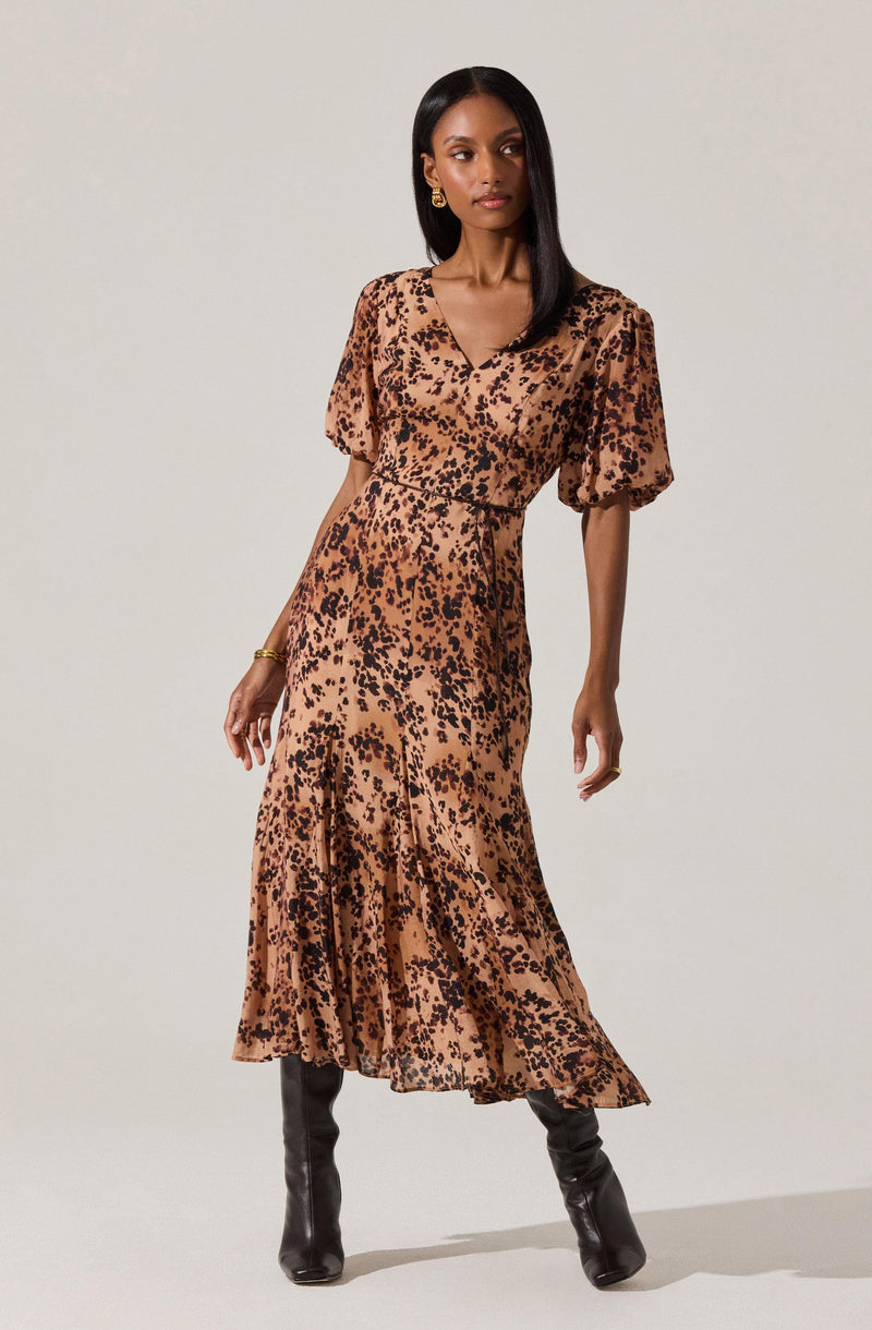 A woman is wearing a midi-length dress with a leopard print pattern in shades of brown and black. The dress features a V-neckline, puffed short sleeves, and a fitted waist. The skirt is flowy with a ruffled hem. She is also wearing black knee-high boots and gold jewelry, including earrings and a bracelet.