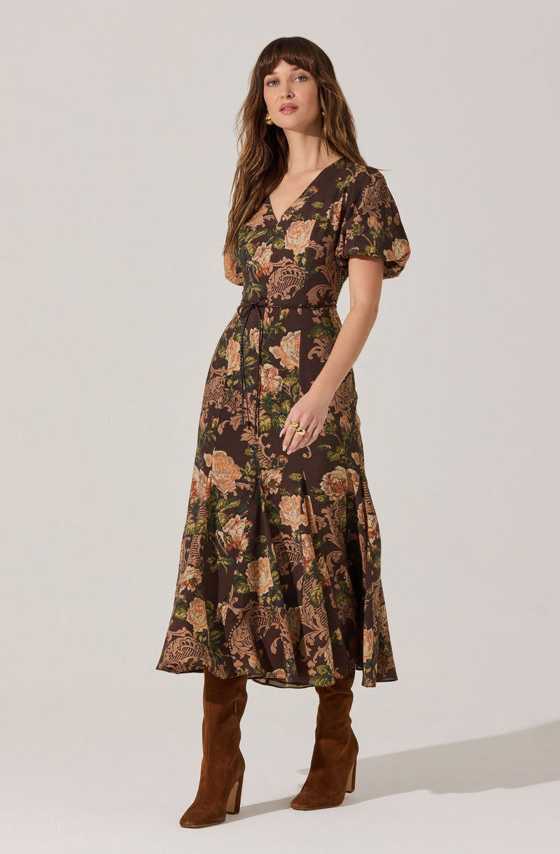 A woman is wearing a long, floral-patterned dress with short, puffed sleeves. The dress features a mix of pink, green, and beige flowers on a dark background, with a V-neckline and a waist tie. She is also wearing high-heeled, brown suede boots.