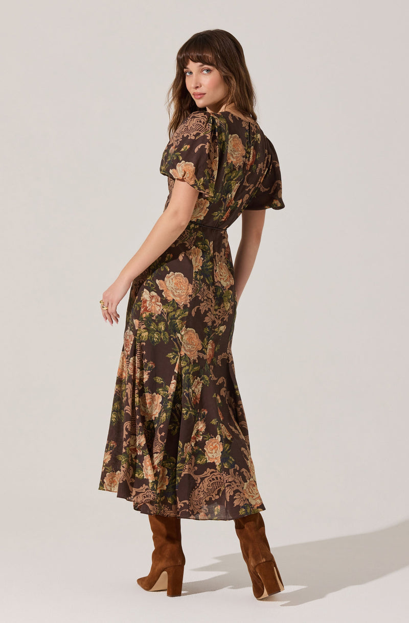 A woman is wearing a long, flowy dress with a dark background and a floral pattern featuring large pink and peach flowers with green leaves. The dress has short, puffed sleeves and a banded waist. The skirt of the dress is slightly pleated and reaches down to her calves, with a subtle slit on the side. She is also wearing brown suede knee-high boots.