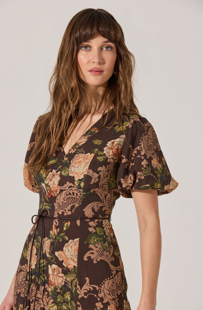 A woman is wearing a dress with a dark brown background featuring large floral patterns in shades of pink, peach, and green. The dress has a V-neckline, short puffed sleeves, and a drawn waist tied with a black cord.