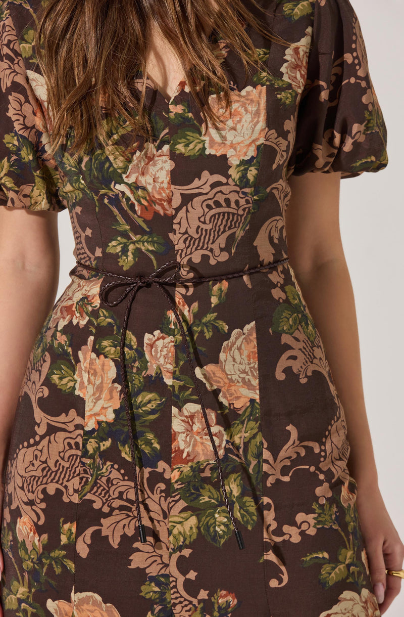 A woman is wearing a dark brown dress with an intricate floral and paisley pattern in shades of pink, peach, green, and beige. The dress has short, slightly puffed sleeves and a waistband with a black drawstring tied in a bow at the front.