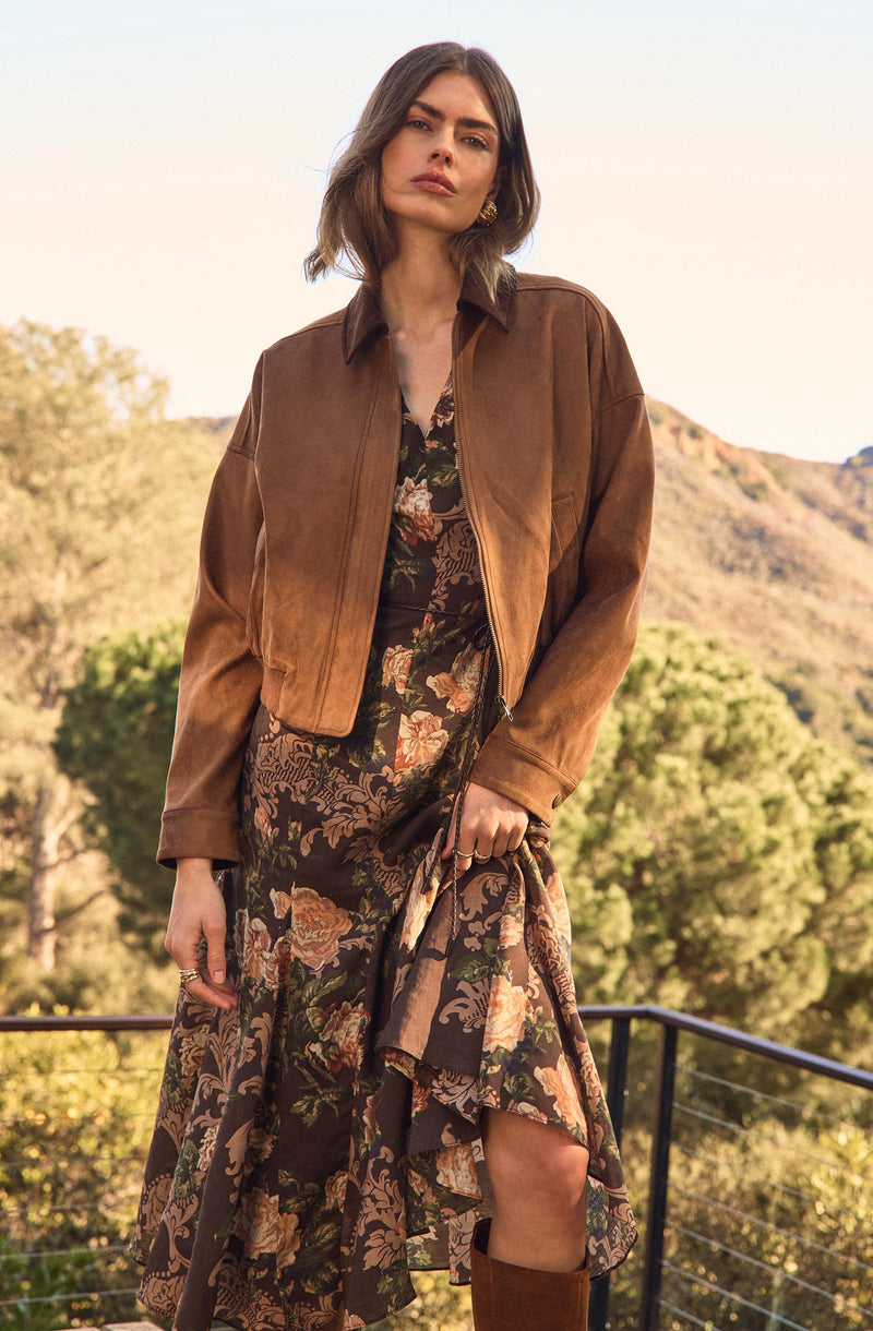 A woman is wearing a brown suede cropped jacket with a collar and long sleeves over a floral dress. The dress features a dark background with a pattern of pink, beige, and green flowers and leaves. She is also holding a portion of the dress, revealing a slit. The woman is accessorized with earrings and rings, and she is standing outdoors with trees and hills in the background.