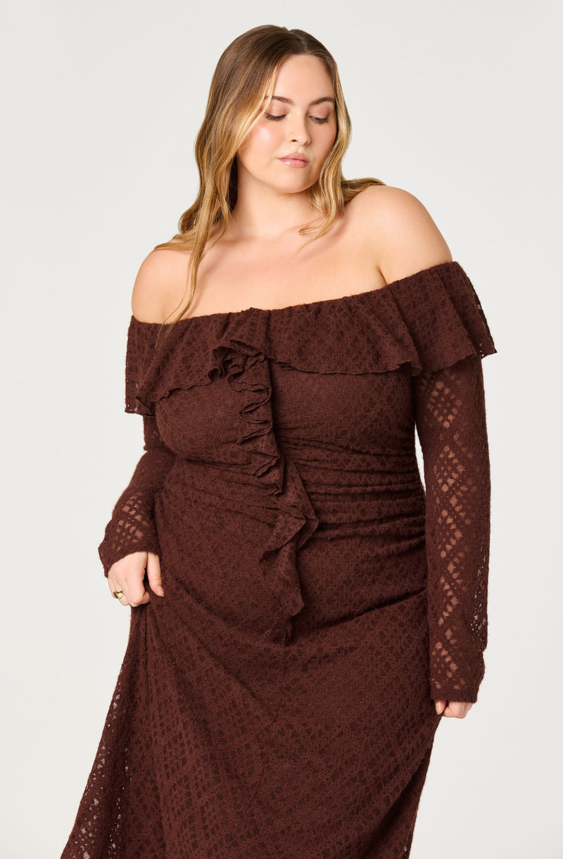 A woman is wearing an off-the-shoulder, long-sleeve dress made of a textured, lace-like fabric in a deep chocolate brown color. The dress features a ruffled neckline that drapes around her shoulders and cascading ruffles down the front.