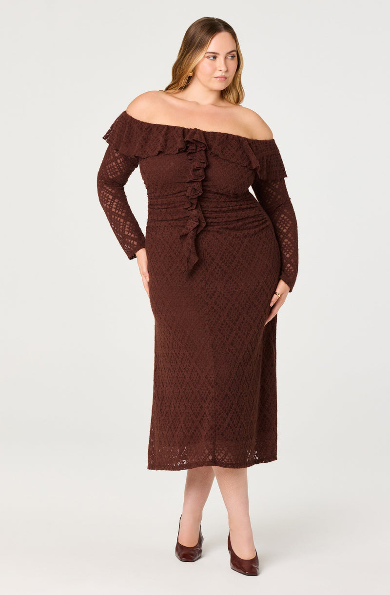 A woman is wearing an off-the-shoulder, long-sleeved, full-length dress made of a textured, lace-like fabric in a rich, dark brown color. The dress features a ruffled neckline that drapes across the shoulders and a vertical ruffle running down the center of the bodice. She is also wearing matching brown high-heeled shoes.