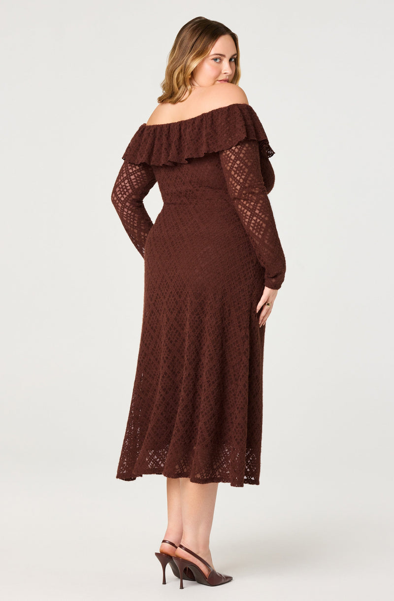 A woman is wearing a floor-length, crochet dress in a deep brown color with long sheer sleeves featuring a diamond-shaped pattern. The dress has an off-the-shoulder neckline with a ruffled overlay that extends around the shoulders and upper arms. She is also wearing brown high-heeled shoes with thin straps.