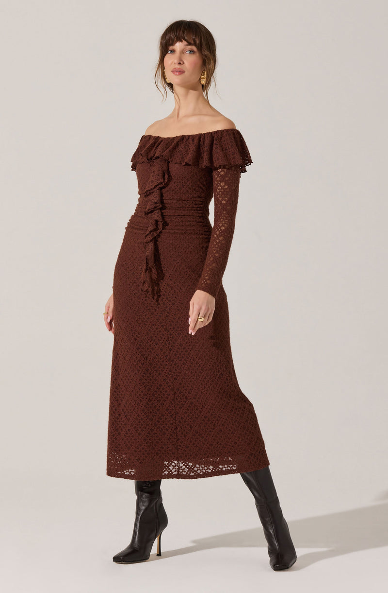 A woman is wearing a long, off-the-shoulder dress in a rich brown color. The dress features a ruffled neckline that drapes over her shoulders and upper arms. It has a textured, lace-like fabric with a delicate pattern, and a fitted waist with ruffle detailing running vertically down the front. The dress extends to her ankles, showing a see-through lace pattern at the hem. She is also wearing knee-high black leather boots with pointed toes and high heels.