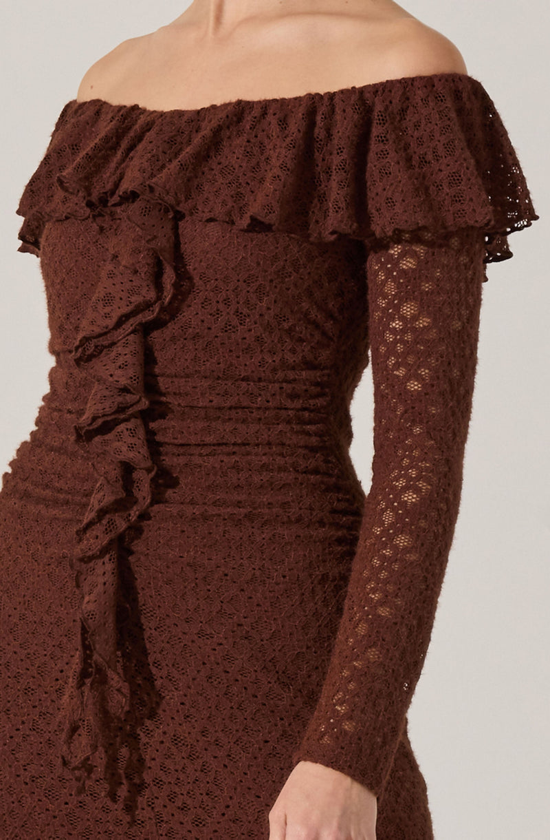 A woman is wearing an elegant off-the-shoulder dress made of brown lace fabric. The dress features a large ruffle that runs along the neckline and extends around the shoulders, adding a decorative touch. The long sleeves are fitted and made from the same lace material, which has a floral pattern. The dress is form-fitting, with ruching details on the sides.