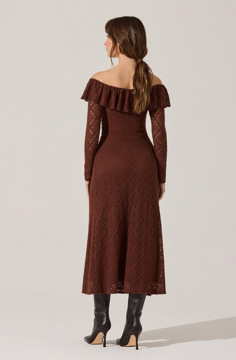 A woman is wearing a long, off-the-shoulder dress in a rich, dark brown color with a textured, lace-like pattern. The dress features a wide ruffle detail around the neckline that drapes over her shoulders and upper arms. The fabric appears semi-sheer with a floral or geometric design, and the dress has a fitted silhouette at the waist that flares slightly towards the hem. She is also wearing tall, black heeled boots.