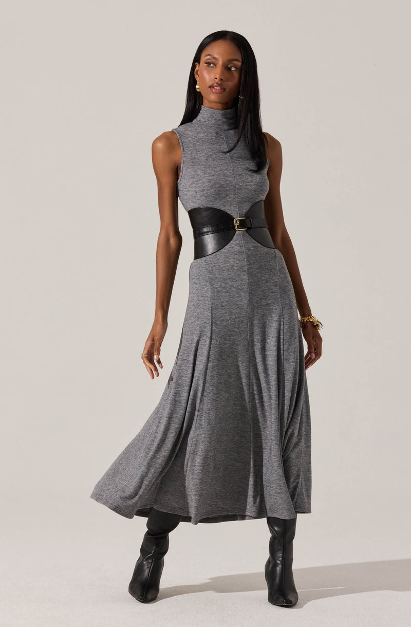 A woman is wearing a sleeveless, high-neck gray dress with a fitted bodice and a flowing, ankle-length skirt. The dress is accented at the waist with a wide black belt that has a gold buckle. She is also wearing black knee-high boots and a gold bracelet on her left wrist.