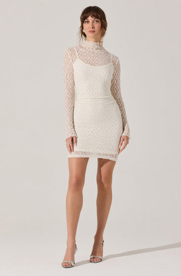 A woman is wearing a white dress with a fitted silhouette. The dress has a high neckline and features a sleeveless design with long, sheer lace sleeves. The lace material has a floral pattern, and the dress combines a lace overlay with a solid lining underneath. She pairs the dress with open-toed high heels.