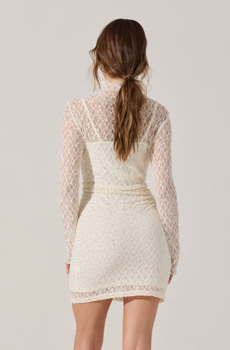 A woman is wearing a long-sleeved, high-neck dress made of white lace with a floral pattern. The dress has a fitted silhouette and falls to mid-thigh length. The lace fabric is see-through on the sleeves and upper back, with a lining on the rest of the dress. The woman’s long, wavy brown hair is styled in a loose ponytail.