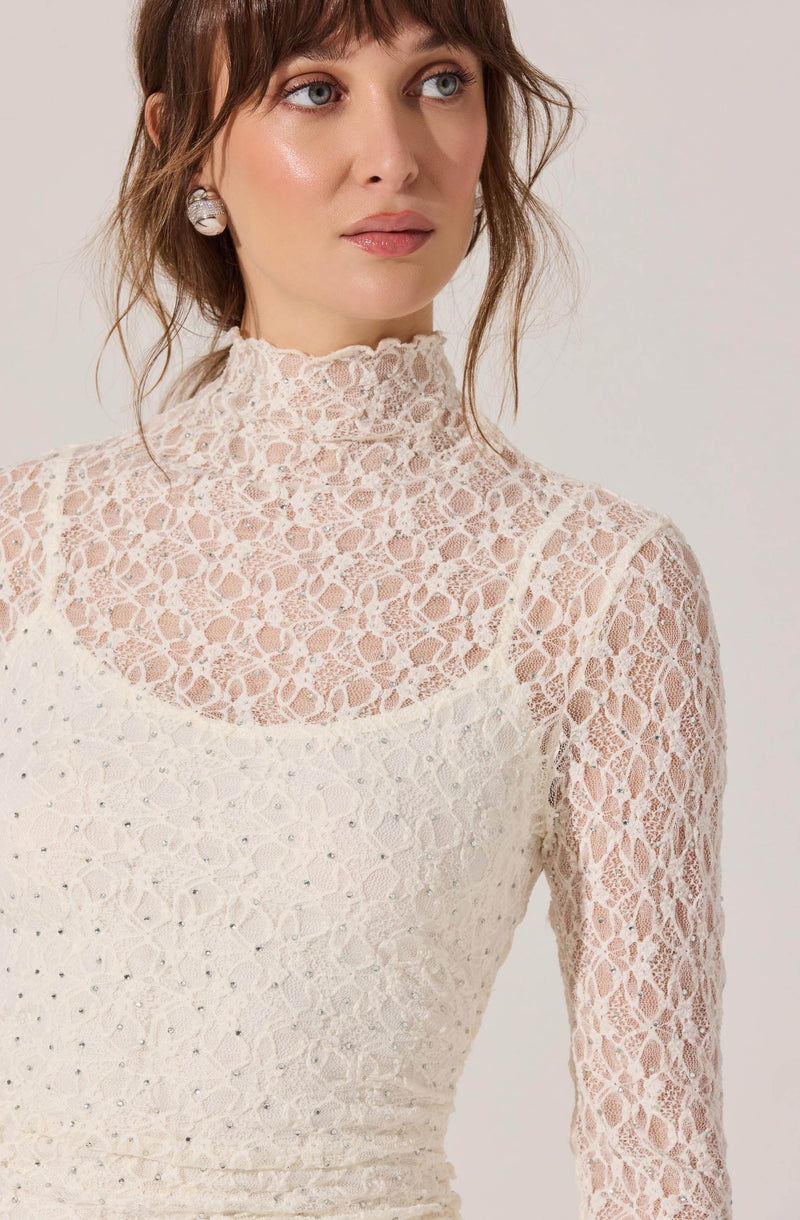 A woman is wearing a high-neck, long-sleeved lace top in a light, natural color. The lace features a floral pattern with intricate detailing, and the top has a fitted silhouette. She is also wearing round earrings.