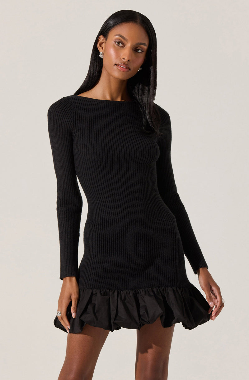 A woman is wearing a fitted, black, long-sleeve, ribbed knit dress with a round neckline. The dress features a ruffled, voluminous hem at the bottom.