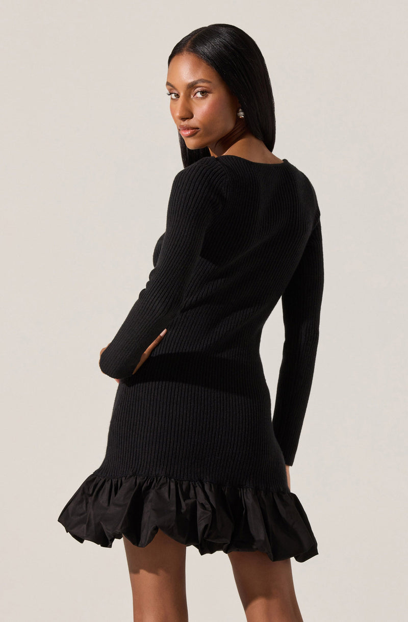 A woman is wearing a black, long-sleeved, ribbed knit dress with a fitted silhouette. The dress features a ruffled hem made from a shiny, gathered fabric.