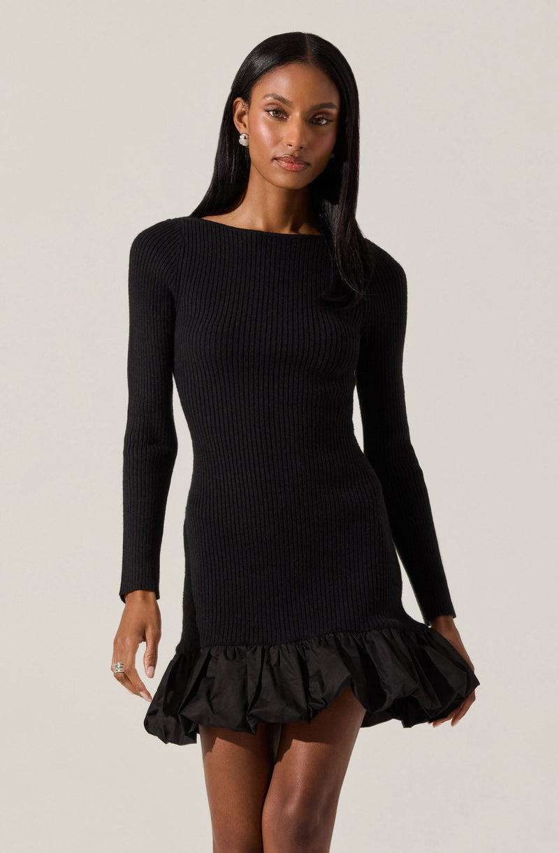 A woman is wearing a black, long-sleeve, ribbed knit dress with a round neckline. The dress is fitted through the torso and hips, and features a ruffled, puffed hem at the bottom that extends above her knees. She has straight, shoulder-length black hair and is accessorizing with large, round earrings.