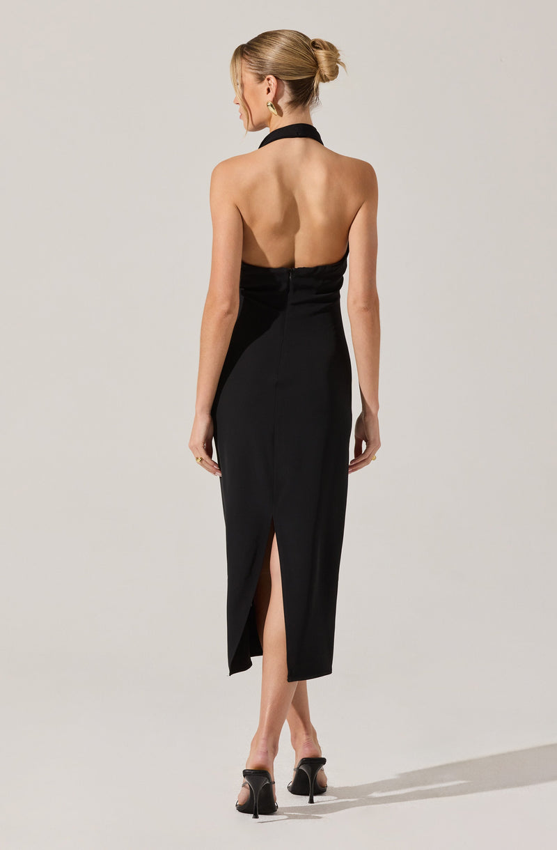 A woman is wearing a long, elegant black dress with a halter neckline and an open back. The dress features a slit at the back, revealing her lower legs. She is also wearing black high-heeled shoes.