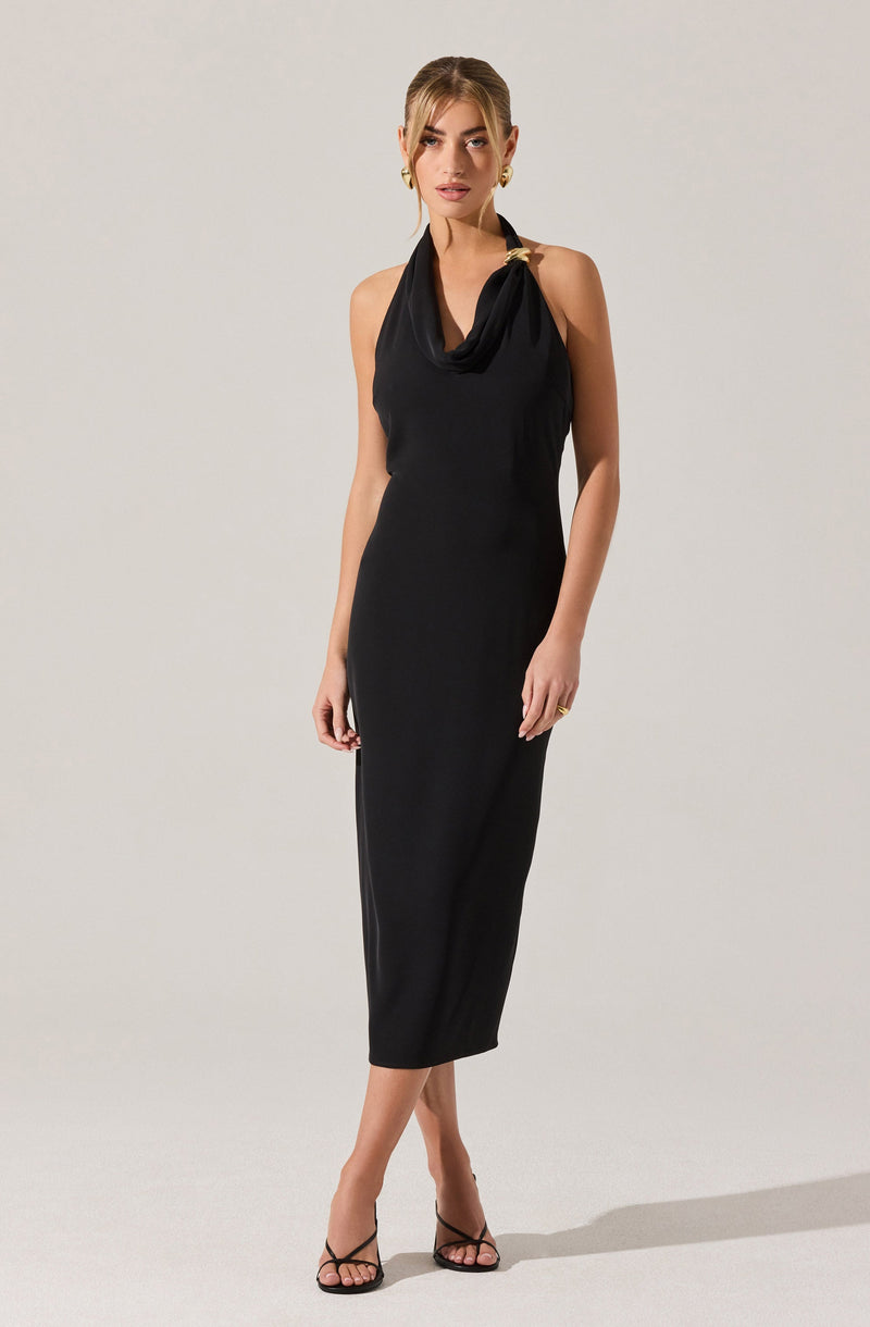 A woman is wearing a sleeveless, black, ankle-length dress with a relaxed fit. The dress features a draped neckline with a gold-tone decorative clasp on one shoulder strap. She is also wearing black strappy high-heeled sandals and gold hoop earrings.