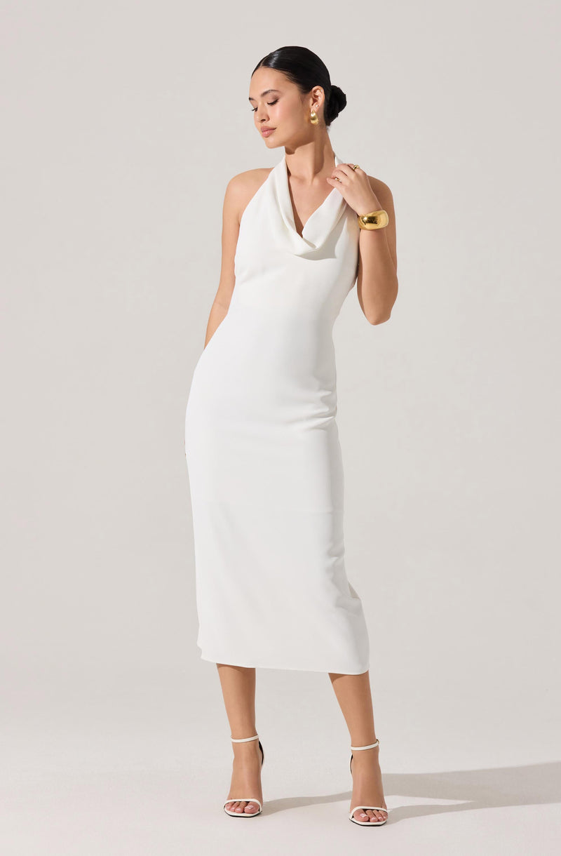 A woman is wearing a sleeveless, white, ankle-length dress with a draped cowl neckline. She is accessorized with gold earrings, a wide gold bracelet, and white high-heeled sandals with ankle straps.
