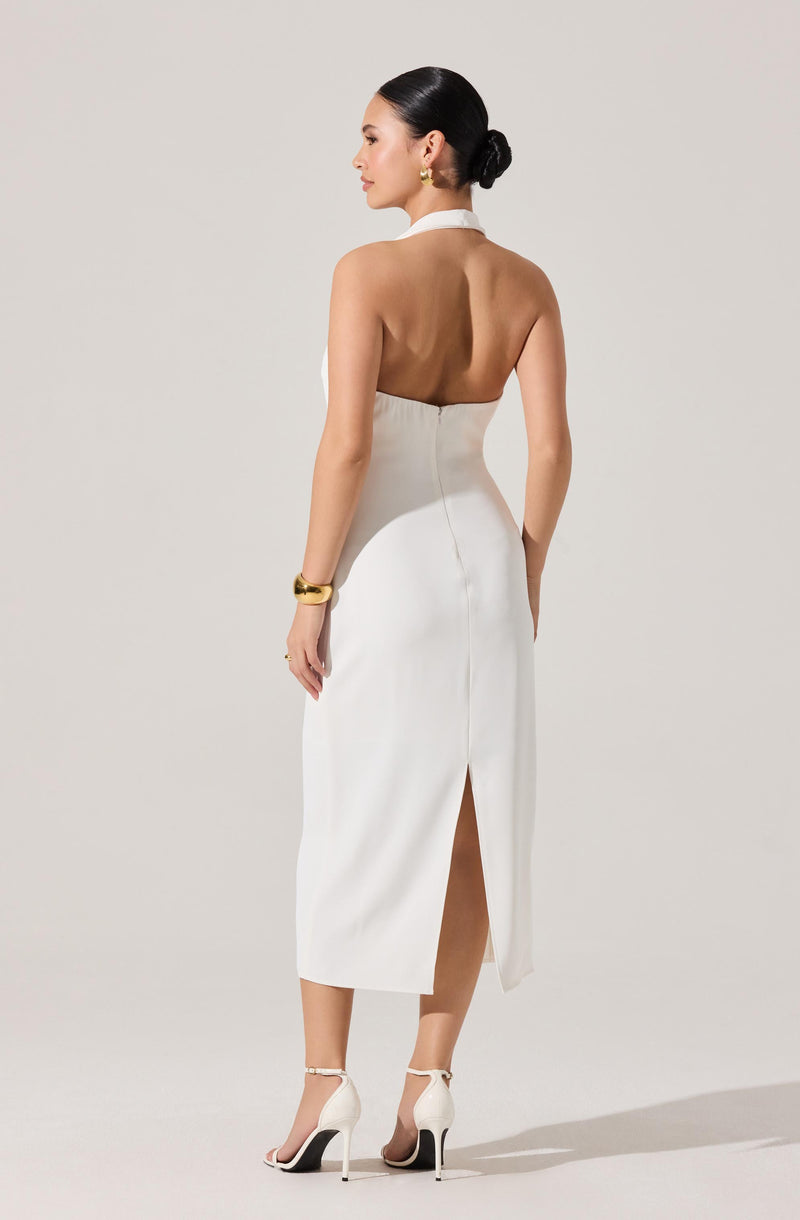 A woman wearing a strapless, backless white dress that extends to mid-calf length, with a slit up the back. She is also wearing white high-heeled shoes with ankle straps, gold earrings, and a gold bracelet on her wrist. Her hair is styled in a sleek, low bun.