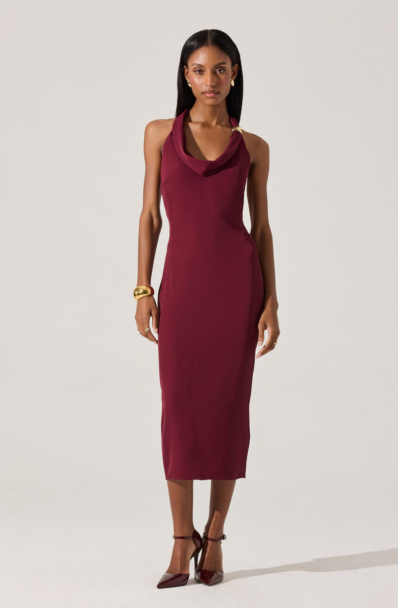 A woman is wearing a sleeveless, form-fitting, ankle-length dress in a deep burgundy color. The dress features a cowl neckline and a gold hardware detail on the left shoulder. She accessorizes with a large gold bracelet on her right wrist and small earrings. She is also wearing matching burgundy high heels with ankle straps.