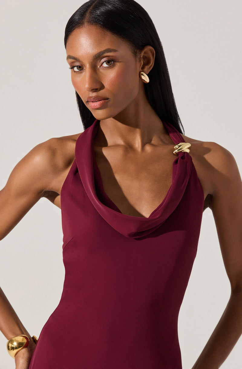 A woman is wearing a sleeveless, deep burgundy dress with a draped cowl neckline. The dress features a decorative gold-tone pin at the shoulder, adding an elegant touch. She has matching gold-tone earrings and a large gold bracelet on her wrist.