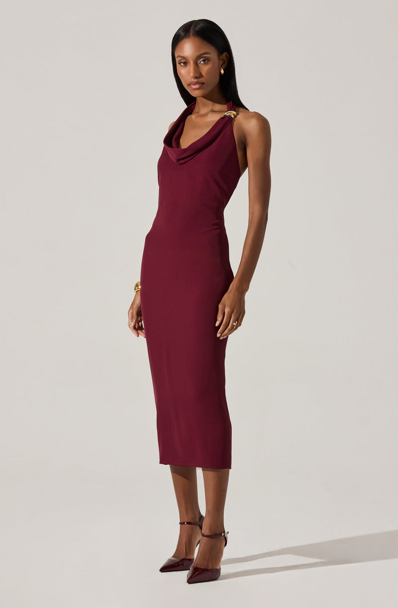 A woman is wearing a sleeveless, form-fitting, mid-length burgundy dress with a draped neckline. The dress features shoulder embellishments with gold hardware. She is also wearing matching burgundy high-heeled shoes and gold jewelry, including earrings and a bracelet.