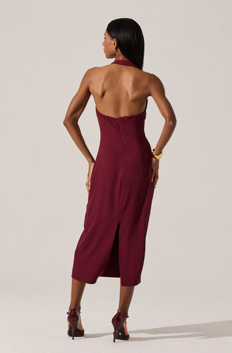 A woman wearing a sleeveless, backless, burgundy halter dress that reaches mid-calf length with a slit in the back. She is styled with matching burgundy high-heeled shoes and gold jewelry, including a bracelet on her right wrist.