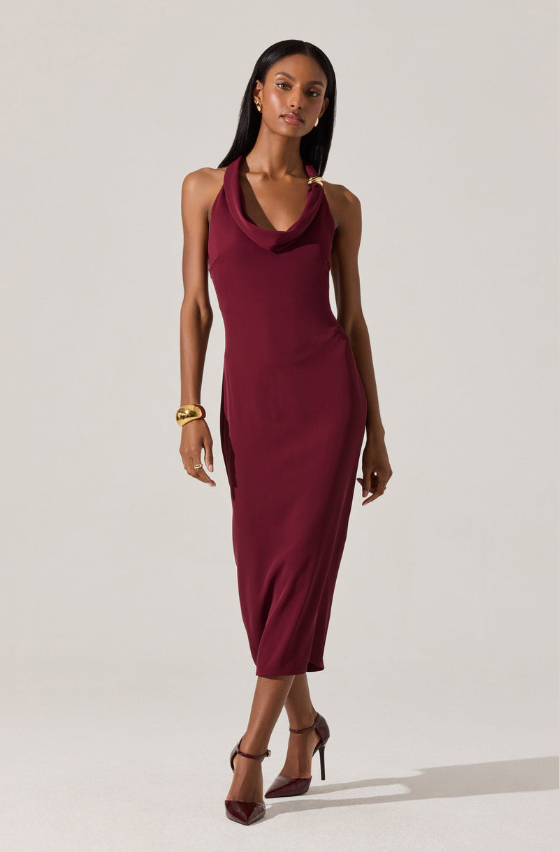 A woman is wearing a sleeveless, ankle-length burgundy dress with a draped neckline. The dress fits closely to the body, emphasizing her shape. She accessorizes with gold jewelry, including a large bracelet on her right wrist and earrings. She is also wearing high-heeled, pointed-toe shoes that match the color of her dress.