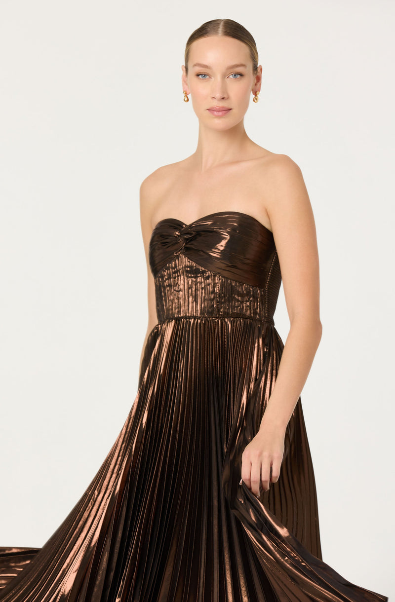 A woman is wearing a strapless, metallic brown gown with a ruched bodice and a pleated, flowing skirt. The fabric has a shiny, reflective quality. She has her hair pulled back and is accessorized with small hoop earrings.