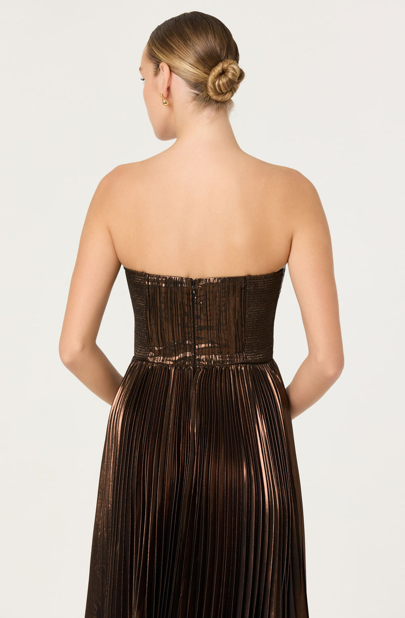 A woman is wearing a strapless dress with a metallic, shiny fabric. The top part of the dress is fitted and appears to have a textured, ruched pattern with a back zipper closure. The skirt part of the dress has vertical pleats and a reflective, coppery or bronze hue. The woman has her hair styled in a low bun and is accessorized with small gold earrings.