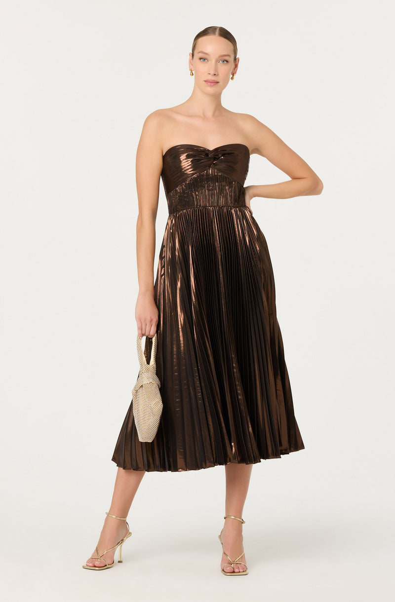 A woman is wearing a strapless, metallic bronze dress with a fitted bodice and a pleated, mid-length skirt. She is holding a small beige textured handbag and wearing beige high-heeled sandals with ankle straps.