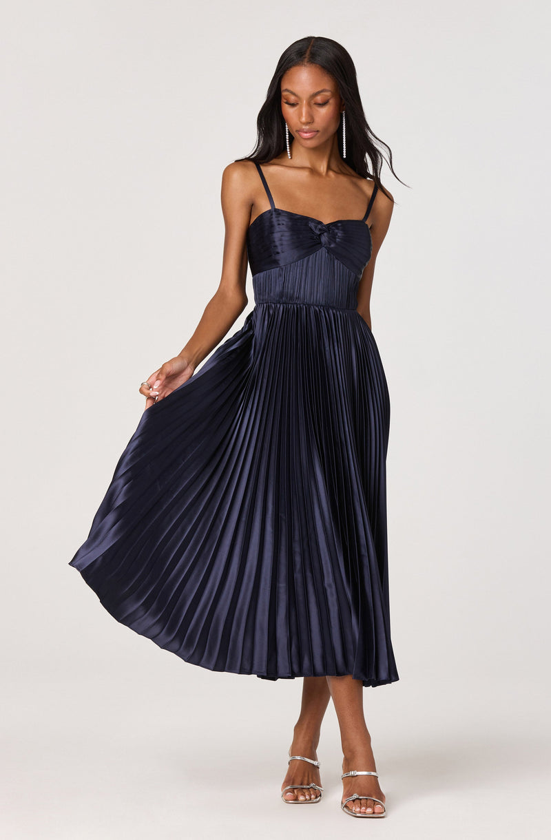A woman is wearing a navy blue dress with spaghetti straps, featuring a fitted, velvet bodice with a bow detail at the chest, and a pleated, satin-like skirt that falls to mid-calf length. She is accessorized with long, dangling earrings and silver high-heeled sandals.