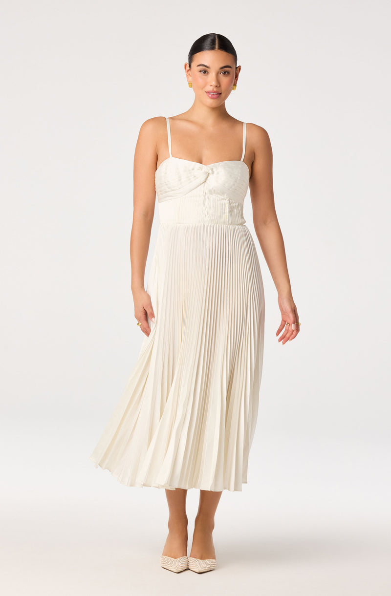 Woman in a cream pleated midi dress with thin spaghetti straps and a bow detail on the bust.
