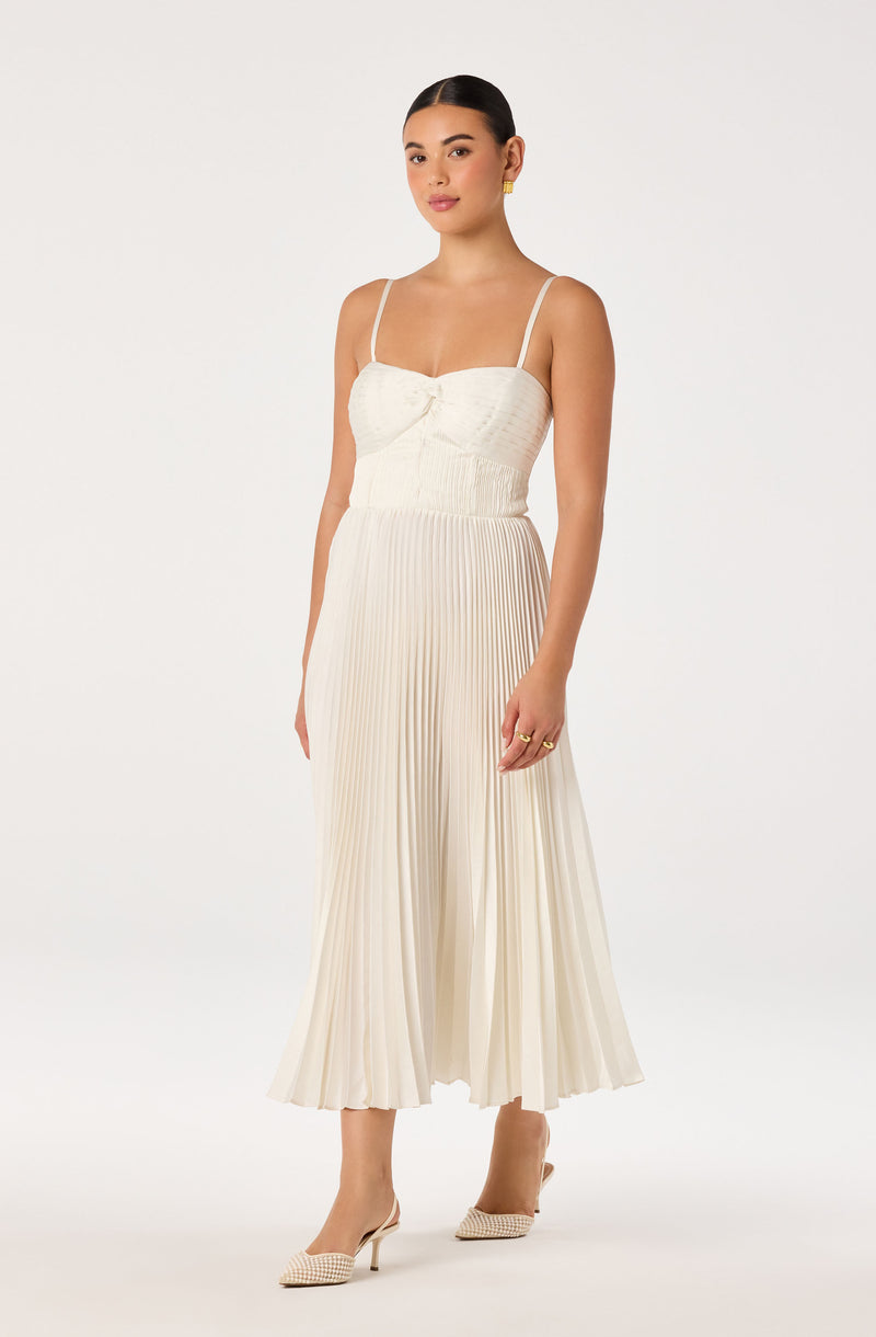 Woman in a cream colored pleated dress with fitted bodice and thin straps, paired with beige heels.
