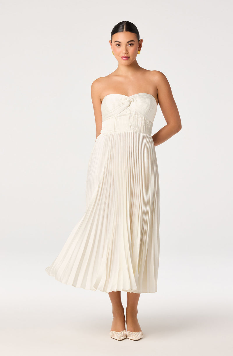 Woman in a strapless, white, pleated dress with a knot detail at the bust, paired with cream heels.