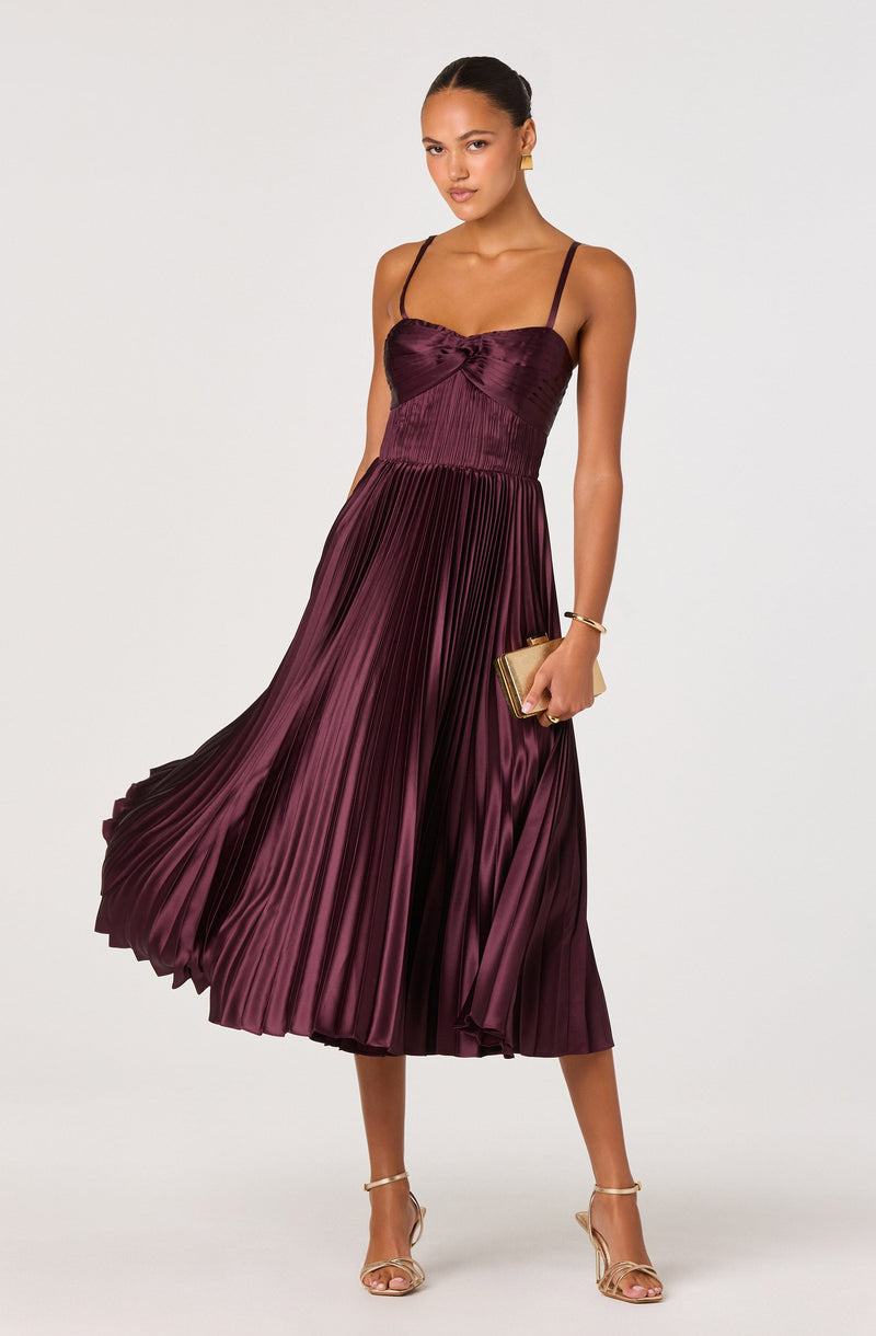 A woman is wearing a sleeveless, burgundy, pleated dress with thin spaghetti straps. The dress features a fitted bodice with a gathered detail at the bust and a flowy, high-waisted skirt that reaches mid-calf length. She is accessorized with gold jewelry, including earrings and a bracelet, and holds a small gold clutch purse. She is wearing nude high-heeled sandals.