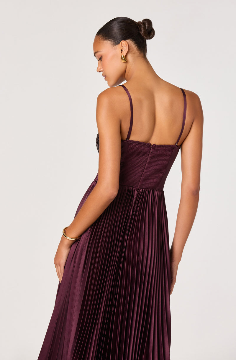 A woman is wearing a sleeveless, deep burgundy dress with thin spaghetti straps. The bodice is fitted with a textured fabric, and the skirt features a pleated design with a smooth, shiny finish. She has her hair styled in a sleek bun and is accessorized with gold hoop earrings and gold bangles.