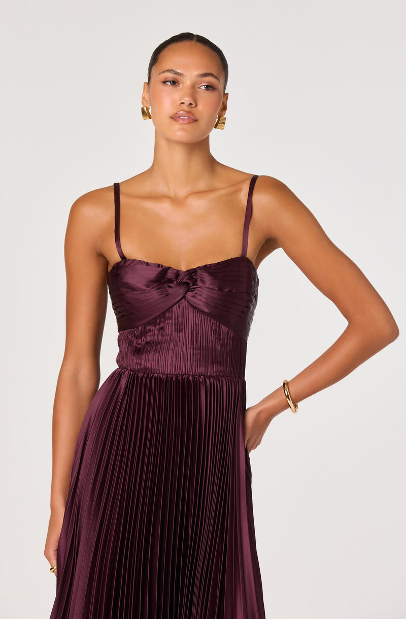 A woman is wearing a sleeveless, deep burgundy dress with thin spaghetti straps. The bodice features a gathered and knotted design, with a slightly shiny, satin-like fabric. The skirt is pleated, creating vertical lines throughout. She has gold hoop earrings and a gold bangle on her wrist.