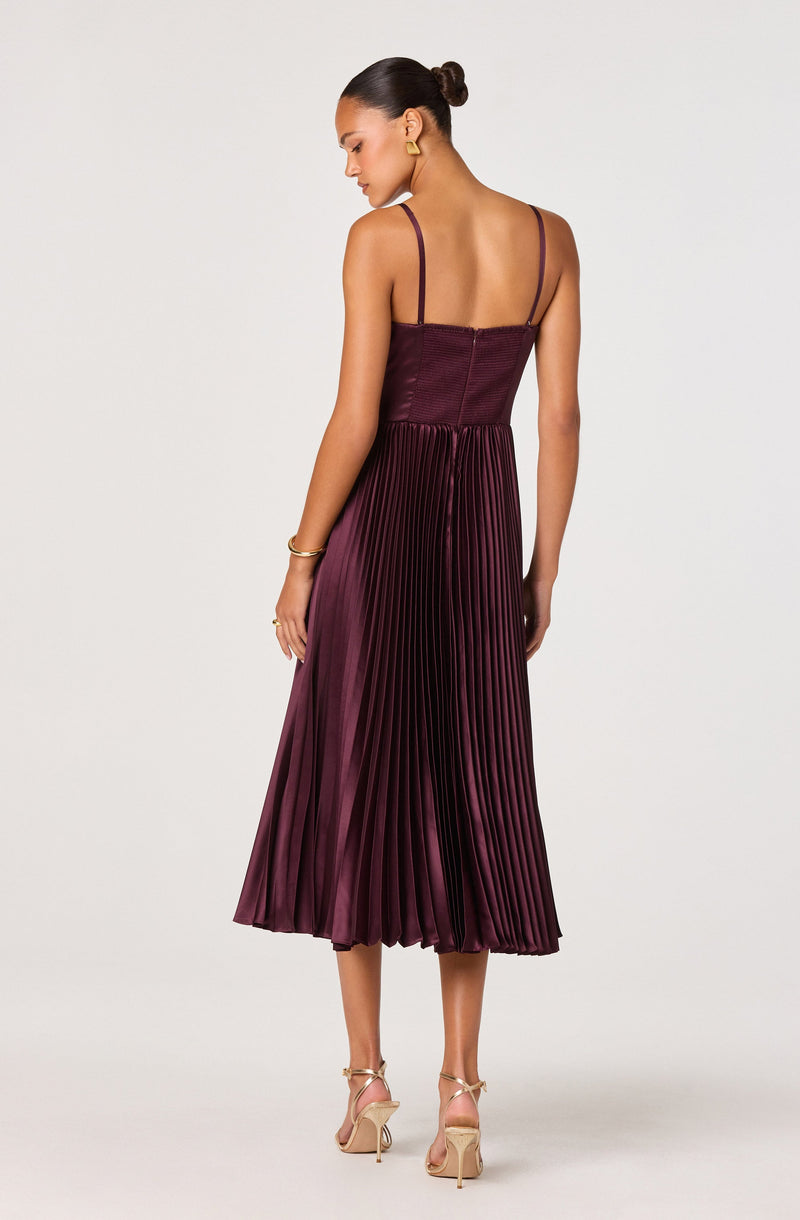 The dress features a fitted bodice with thin spaghetti straps and a zipper closure at the back. The skirt is pleated, with a shiny satin-like fabric in a deep burgundy color, creating a flowing and elegant appearance.