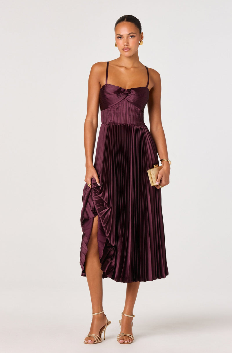 The dress is a burgundy, satin fabric with a fitted, ruched bodice and thin spaghetti straps. The skirt features a high-low hemline with pleats and a ruffled detail on one side, creating a side slit that reveals part of the leg. The dress has a shiny, elegant appearance suitable for formal occasions.