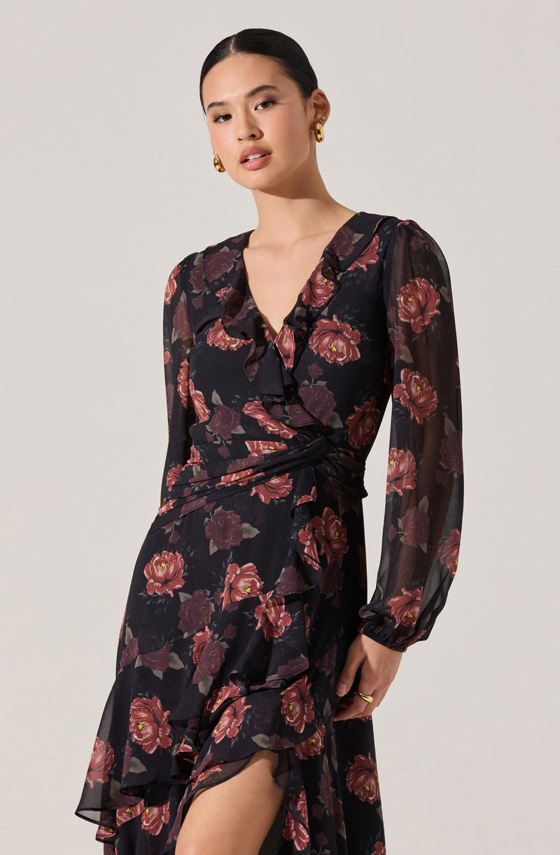 A woman is wearing a long, flowing dress made of sheer fabric with a black background and a large floral print in shades of red, pink, and burgundy. The dress features long, semi-transparent puffed sleeves, a V-neckline with ruffled details, and a ruffle along the hemline. The waist is cinched, creating a feminine silhouette, and the dress has a high slit revealing part of her leg. She accessorizes with gold hoop earrings and a ring.