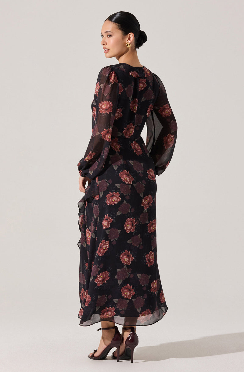 A woman is wearing a long, semi-sheer black dress with a red and dark purple floral pattern. The dress features long, slightly puffed sleeves and a round neckline. She is also wearing high-heeled sandals and gold earrings, with her hair styled in a sleek, low bun.